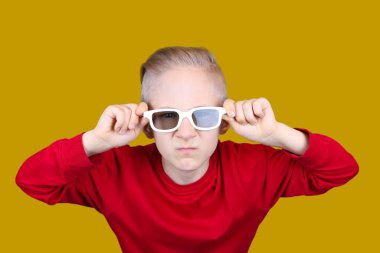 Kırmızı ceketli bir çocuk sarı arka planda ellerini gözlüğünün arkasında tutuyor.