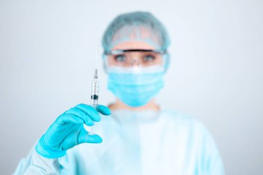 Tıbbi elbise, maske ve koruyucu eldiven takan şeffaf gözlüklü bir hemşire elinde bir şırınga tutuyor.