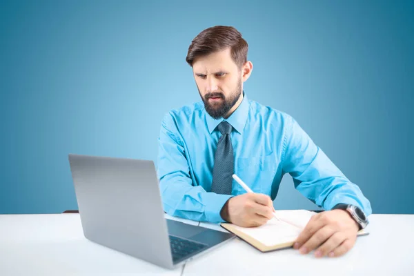 ノートパソコンに座っている青いシャツを着たビジネスマンが 顔にリラックスした表情でノートパソコンにタスクを書き込みます — ストック写真