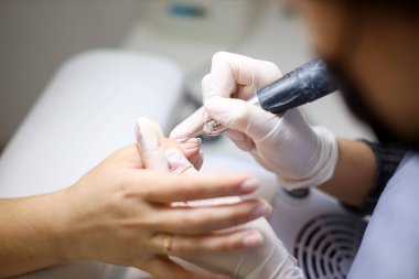 Manikür ustası, müşterinin tırnaklarını törpüleme makinesiyle kesiyor. Elinde bir aletle manikürcü tırnaklarını gıcırdatıyor ve oje sürmeye hazırlanıyor.