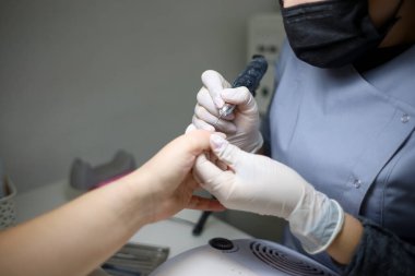 Manikür ustası müşterinin parmaklarını tutuyor ve manikür makinesiyle inceliyor.