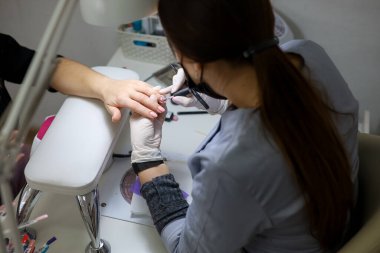 Kız manikür ustası manikür yapıyor. Mavi gömlekli ve siyah maskeli bir manikürcü müşterinin tırnaklarını yapar.