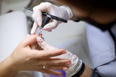 Manikürcü, tırnak cilası sürerek müşteriye manikür yaptırıyor. Beyaz eldivenli bir manikür ustası müşterinin tırnaklarıyla çalışır.