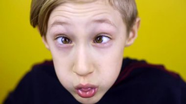 Çocuk gözlerini burnuna koyar ve komik yüzler gösterir. Çocuk sarı arka planda duruyor ve duygularını gösteriyor.