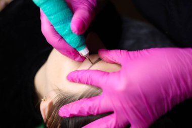 Kalıcı bir makyaj sanatçısı, dövme prosedürünü uygulamak için kaşların yakınındaki deriyi sıkılaştırır. Gözlerinin yakınındaki eldivenler ve deri pigmentle dağılmış.