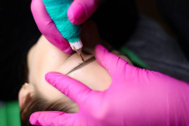 Kalıcı bir makyaj sanatçısı, dövme prosedürünü uygulamak için kaşların yakınındaki deriyi sıkılaştırır. Gözlerinin yakınındaki eldivenler ve deri pigmentle dağılmış.