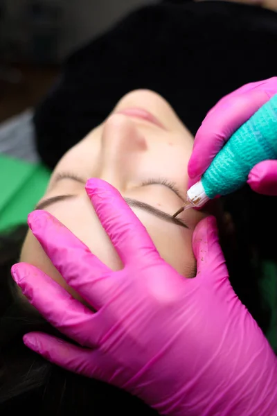 The master performs eyebrow tattooing. The gloves and the skin near the eyes are splattered with pigment