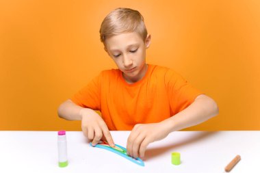 Turuncu tişörtlü bir çocuk beyaz bir masaya oturur ve ince şeritli kağıtları yapıştırmak için hazırlar. Çocuk tüy dökme tekniğini öğrenir.