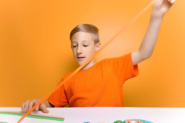 Turuncu tişörtlü bir çocuk önünde ince bir kağıt şeridi uzatıyor.