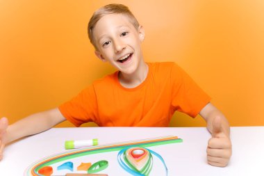 Turuncu tişörtlü bir çocuk tüy dökme tekniğindeki ince kağıt şeritleri bükmeden önce kameraya güler.