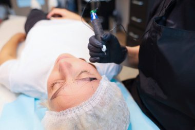 Müşteri kalıcı kaş makyajı prosedürünün altında yatıyor ve yüzünde bir çizim var ve dövme sanatçısı prosedürü gerçekleştiriyor..