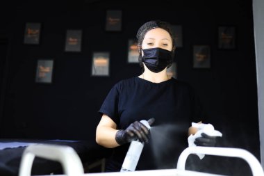 Dövmeci iş yüzeyini dezenfekte eder, kalıcı makyaj için hazırlık çalışması yapar. Siyah maskeli dövmeci kız önünde dezenfektan testeresi kullanıyor