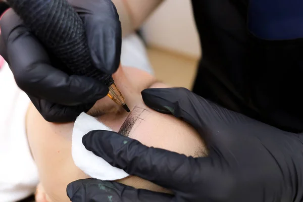 the master of permanent makeup performs a tattoo of the eyebrows with a tattoo machine holding the eye area with a cotton sponge