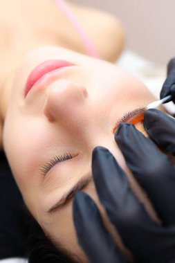 Kız model kozmetik kanepede yatıyor. Silia kaplamasının ustasının elleri. Silia 'yı mankenin göz kapağına kıvırmak için silindiri bastırıyor.