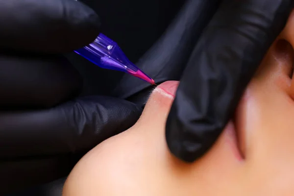 a tattoo artist in black gloves holds a tattoo machine, and the other finger presses on the client\'s lips, thereby performing a lip tattoo