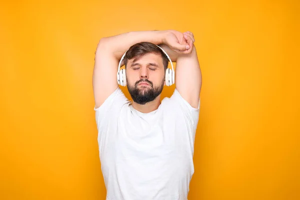 a bearded man wearing white headphones, closing his eyes, and listening to calm music, throwing his hands behind his head.