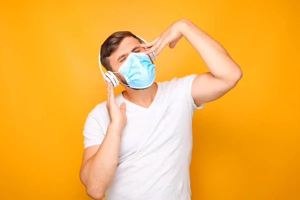 a man in white musical headphones stands on a yellow background, wearing a medical mask.