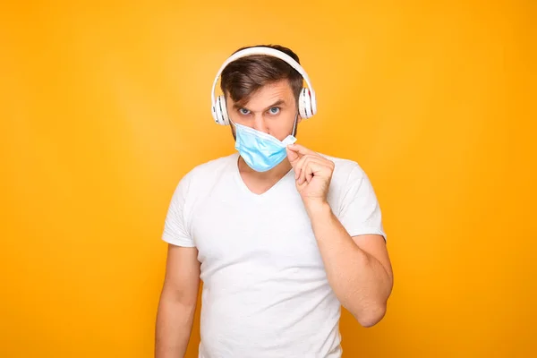 a man with musical headphones and a medical mask stands against a yellow background.
