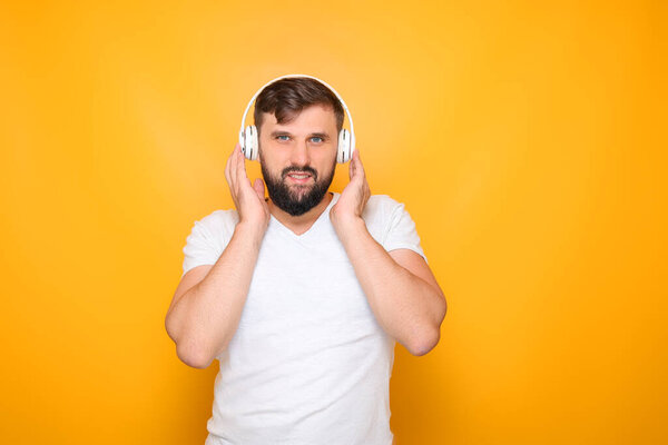a bearded man standing against a yellow background presses white headphones to his ears and listens to music.