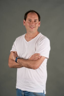 Kollarını gri bir arka plana çarpmış genç bir adamın portresi. Beyaz tişörtlü ve mavi saatli sıradan ve profesyonel tarzda. Temiz ve çok yönlü resim.
