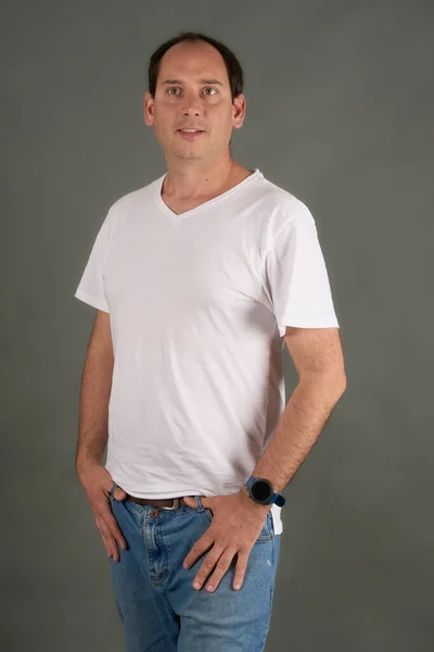 Kel bir adamın portresi, elleri kalçasında, gri bir arka planda. Beyaz tişörtlü, rahat ve kendinden emin bir tarz. Ticari kullanım için temiz ve çok yönlü resim.