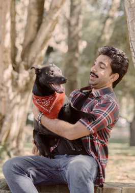 İnsan ve köpek orman parkında huzurlu bir anın tadını çıkarırlar. Kırmızı bandanalı siyah köpek tetikte görünüyor. Ağaçların arasından gün ışığı, duygusal bağ, yaşam tarzı ve evcil hayvan fotoğrafçılığı.