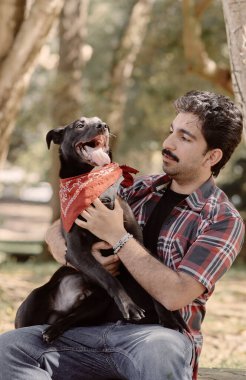 İnsan ve köpek ormanda sakin bir anı paylaşır. Kırmızı pantalonlu siyah köpek. Sahibi evcil hayvana sevgiyle bakar. Doğal ışık, yaşam tarzı ve evcil hayvan fotoğrafçılığı.