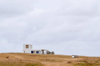 Cabo Polonio, Uruguay 'da modern bir ev. Büyük pencereleri ve kırmızı çatısı olan minimalist beyaz bir yapı, uzak bir kıyı manzarasında bulutlu bir gökyüzünün altında çimenli bir tepede kurulmuş..
