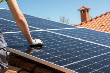 Güvenlik donanımlı bir işçi, parlak mavi gökyüzünün altındaki bir çatıya dikkatlice güneş panelleri yerleştiriyor. Görev, verimlilik ve istikrarı sağlamak için panelleri sıkıca güvence altına almayı içeriyor..