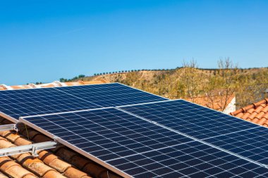 Güneş panelleri açık mavi bir gökyüzünün altında fayanslı bir çatıya monte edilir. Kurulumda konut kullanımı için çevre dostu enerji üretimi sergileniyor.