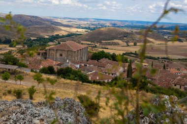 Guadalajara 'daki Atienza' ya bakan bu bakış açısı, bulutlu bir gökyüzünün altındaki yuvarlak tepeler ve yeşillikler tarafından çerçevelenmiş geniş bir kırsal alana kurulmuş tarihi binaları gözler önüne seriyor..