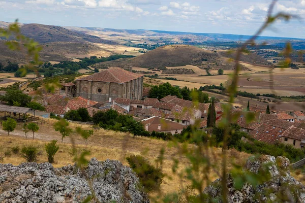 Guadalajara 'daki Atienza' ya bakan bu bakış açısı, bulutlu bir gökyüzünün altındaki yuvarlak tepeler ve yeşillikler tarafından çerçevelenmiş geniş bir kırsal alana kurulmuş tarihi binaları gözler önüne seriyor..
