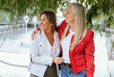 İki kadın bir arada durur, gülümser ve kucaklaşır, dışarıda çeşmenin yanında vakit geçirirken. Bahçe yemyeşil ve yemyeşil, neşeli bir atmosfer yaratıyor..