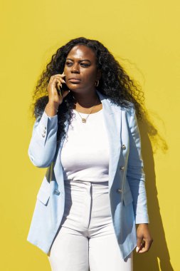 Bir kadın açık mavi bir ceket ve beyaz bir bluzla sıcak bir şekilde gülümsüyor. Canlı sarı bir duvarın önünde poz veriyor güneşli günün tadını çıkarıyor ve tarzını sergiliyor..