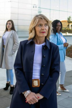 Çeşitli stillere sahip üç kadın şık bir ofis binasının dışında güven içinde poz veriyor. İş kıyafetlerinde güç ve dostluk gösterirken güneş parlıyor..