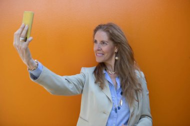 Canlı turuncu bir duvarın önünde duran bir kadın selfie çekmek için akıllı telefonunu tutuyor. Uzun, dalgalı saçları var ve açık renk ceket ve mavi gömlek giyiyor..