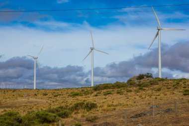 Yükselen rüzgar türbinleriyle bezenmiş yuvarlanan tepeler boyunca hafif bir esinti esiyor. Geniş bulutlara karşı kuruluyor. Bu barışçıl manzara doğanın enerjiyle uyumunu gösteriyor..