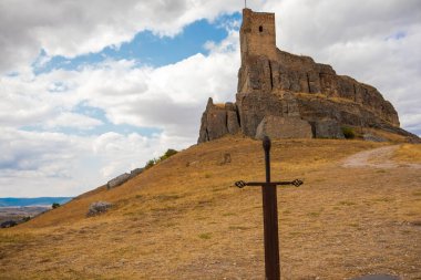 Ziyaretçiler, İspanya 'nın Guadalajara kentindeki Atienza kayalık bir tepenin üzerinde, ön planda bulutlu bir gökyüzünün altında göze çarpan bir kılıç anıtı bulunan çarpıcı bir kaleyi keşfediyorlar..