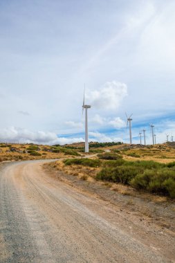 Parlak, kısmen bulutlu bir gökyüzü altında sürdürülebilir enerji üretimi gösteren kayalık çıkıntılı çayırların üzerinde birkaç rüzgar türbini yükseliyor..