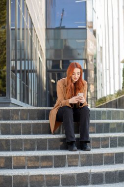 Kızıl saçlı genç bir kadın taş basamaklarda oturuyor, telefonuna odaklanmış. Modern bina güneş ışığını yansıtıyor, canlı bir şehir atmosferi yaratıyor..