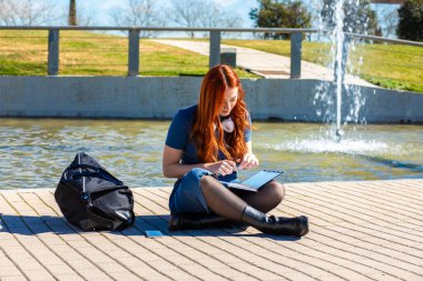 Canlı bir park ortamında, genç bir kadın fıskiyenin yanında oturur ve derslerine odaklanır. Güneş parlıyor, notlar yazarken huzurlu bir atmosfer yaratıyor..