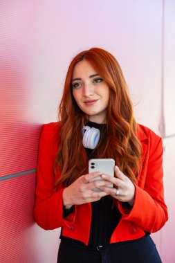 Uzun kızıl saçlı genç bir kadın pembe bir duvarın karşısında duruyor. Akıllı telefonuna odaklanırken parlak kırmızı bir ceket ve kulaklık giyiyor. Sahne çağdaş ve canlı görünüyor..