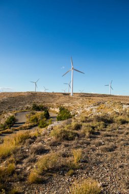 Rüzgâr türbinleri, kuru çimenler ve kayalık arazilerle çevrili sakin bir arazide uzun süre ayakta kalırlar. Ayar doğal ortamda yenilenebilir enerjinin güzelliğini gösteriyor..