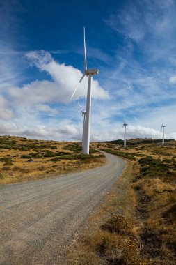 Uzun rüzgar türbinleri yuvarlanan tepelerle çevrili güzel bir manzarada dururlar. Yol, parlak mavi gökyüzünün ve kabarık beyaz bulutların altında gezinerek yenilenebilir enerji sergiliyor..