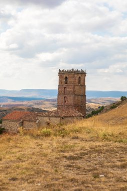 İspanya 'nın Guadalajara kentindeki Atienza şehrinde, arka planda parçalı bulutlu bir gökyüzünün altında uzanan geniş tepeler ve tarlalar ile çarpıcı bir tarihi kule ayakta duruyor..