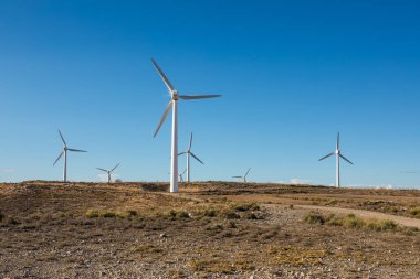 Uzun rüzgar türbinleri açık mavi bir gökyüzünün altında geniş bir çöl arazisine dağılmış durumda. Bu sahne yenilenebilir enerji ve sürdürülebilirliğin önemini vurgular..