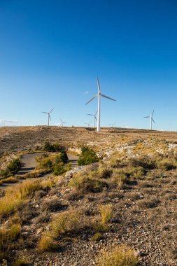 Rüzgâr türbinleri, kuru çimenler ve kayalık arazilerle çevrili sakin bir arazide uzun süre ayakta kalırlar. Ayar doğal ortamda yenilenebilir enerjinin güzelliğini gösteriyor..