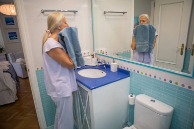 Açık pijamalı bir kadın neşeli bir banyoda duruyor, yüzünü mavi bir havluyla kurutuyor. Mekan ışıkla dolu, sakin bir sabah atmosferini yansıtıyor..