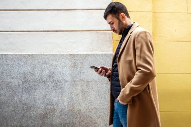 Şık montlu genç bir adam, bir şehir sokağında canlı bir duvara yaslanmış duruyor. Akıllı telefonuna odaklanmış, bir mesaja dalmış gibi, düşünceli bir ifadeyle..