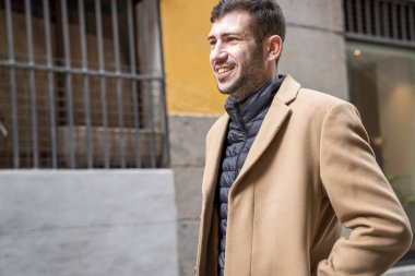 Neşeli bir adam sıcak bir palto giymiş, canlı bir caddede yürüyor. Güneş ışığı etrafındaki modern binaları vurguluyor. Rahatlamış görünüyor, şehrin canlı atmosferinin tadını çıkarıyor..